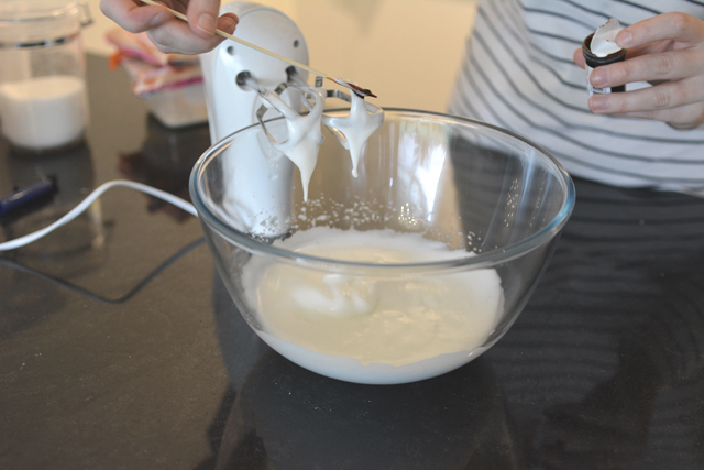 an image of a bowl filled with a frothy mixture, with a pair of woman's hands gently adding food coloring to the mixture. a mixer rests nearby, positioned beside the bowl
