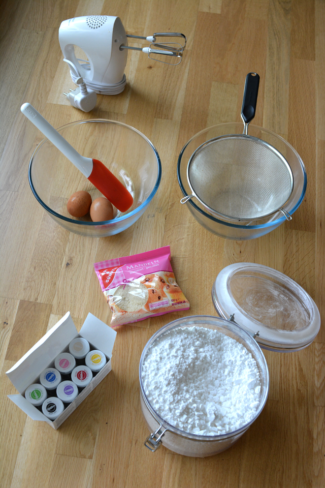 top-down view of french macarons ingredients and equipment: a bowl with 2 eggs and a spatula, a bowl with a strainer, a box of food coloring, a canister of flour, a pack of icing sugar, and a hand mixer