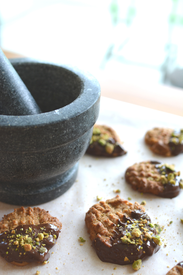 flourless peanut butter cookies, dipped in chocolate and sprinkled with pistachios. arranged flat on parchment paper, with a stone and mortar and pestle beside them