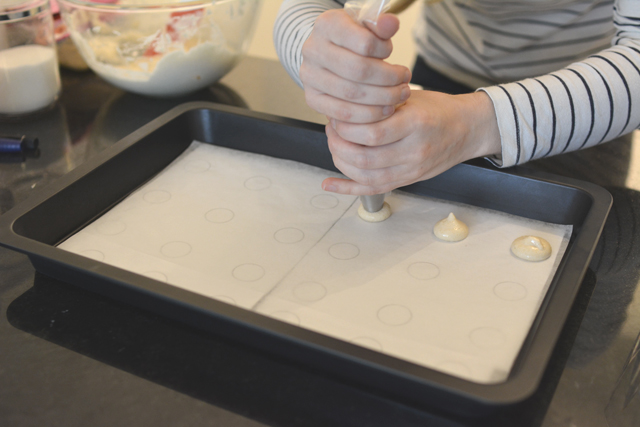 a pair of woman's hands delicately piping the macaron mixture into a template on a baking tray
