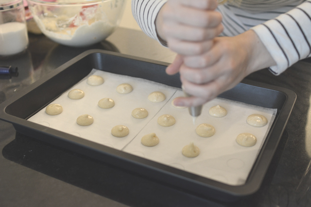 close up image of a pair of woman's hands delicately piping the macaron mixture into a template on a baking tray
