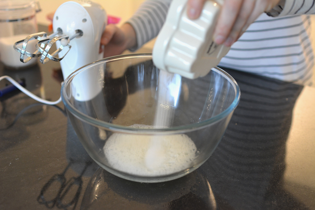 image of a bowl with egg whites, a woman's hand pouring sugar into the bowl, while her other hand is holding a hand mixer