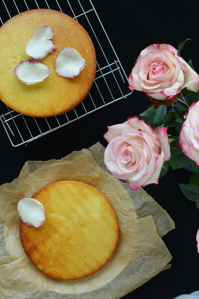 an image of a two-layer cake: one layer is resting on a cooling rack, and the other layer is on parchment paper. the cake layers are adorned with delicate rose petals, and three fresh rose flowers are placed nearby