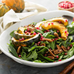 A white bowl filled with roasted acorn squash, spinach, and pomegranates with Acorn squash in the background.