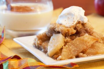 A plate of apple crisp with a dollop of whipped cream and some eggnog in the background.