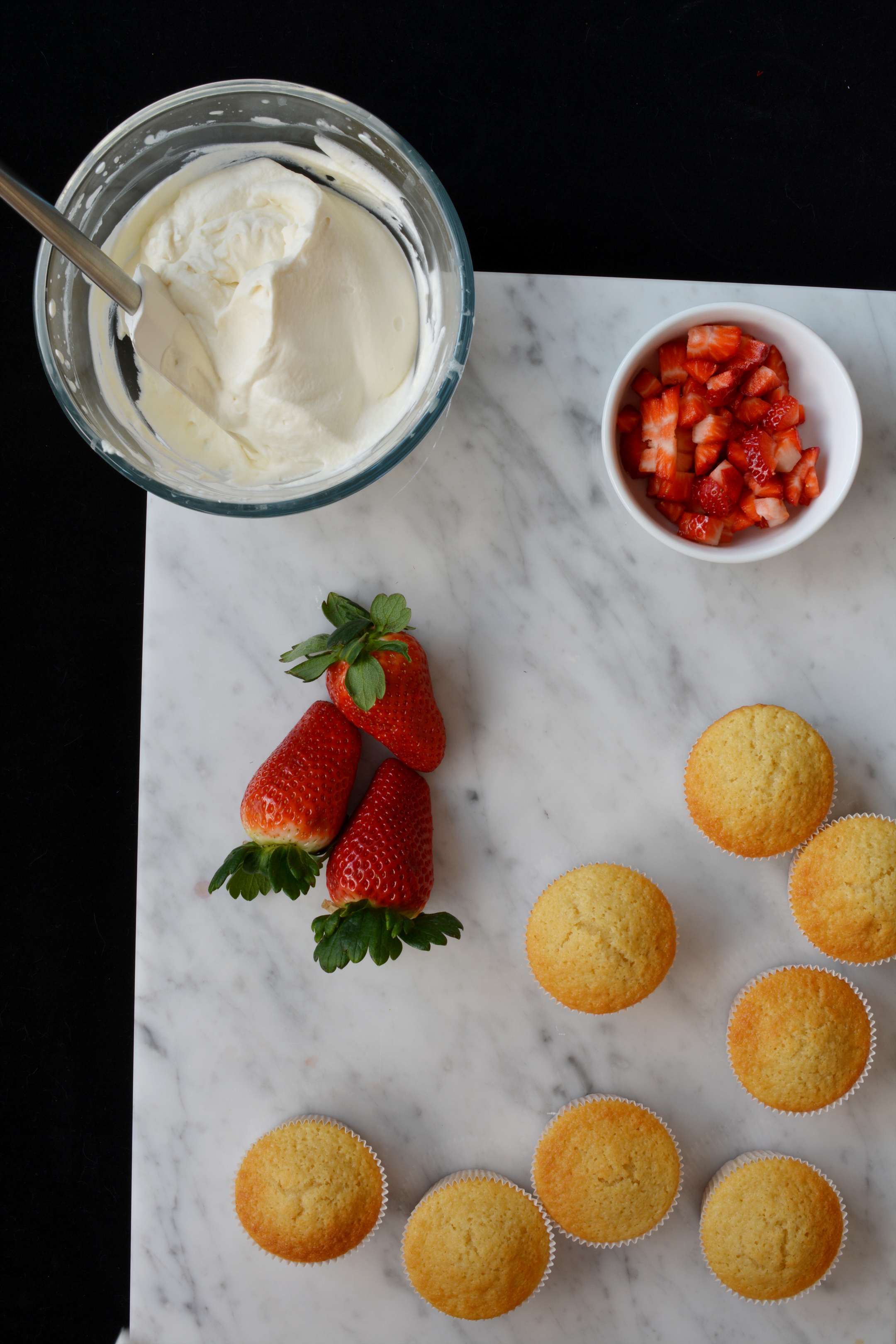 top view of freshly baked cupcakes in a batch, alongside a bowl of cubed strawberries and a separate bowl containing a cream mixture, with fresh strawberries placed nearby