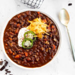 Black Bean Chili with Chocolate in a white bowl garnished with jalapeno slices, sour cream, and shredded cheddar, top view.