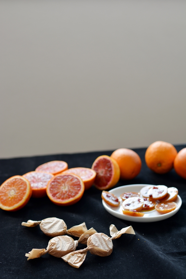 plate of salted blood orange toffees with fresh orange slices and whole oranges, accompanied by three individually wrapped toffees resembling candies