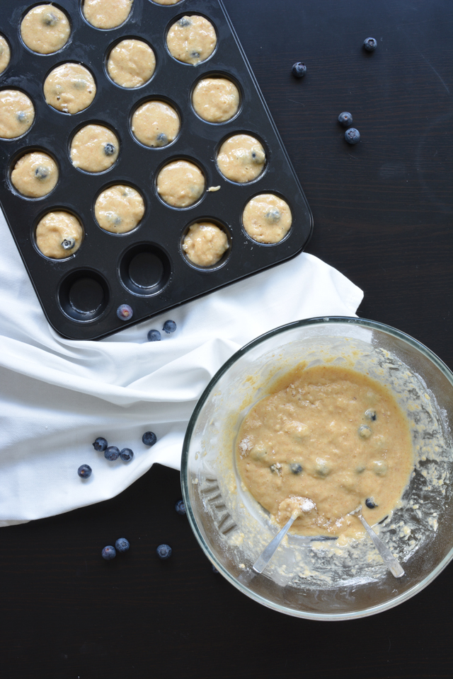 an overhead view of a muffin tray filled with muffin batter and topped with blueberries, accompanied by a bowl containing more muffin batter next to it
