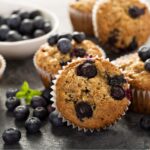 Blueberry muffins on a dark surface with more muffins and blueberries in the background.