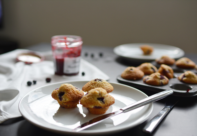 close-up photograph of a plate featuring three mini muffins of brown butter and blueberries, alongside a fork. in the background, there is a muffin tray filled with freshly baked muffins and a jar of jam placed next to it
