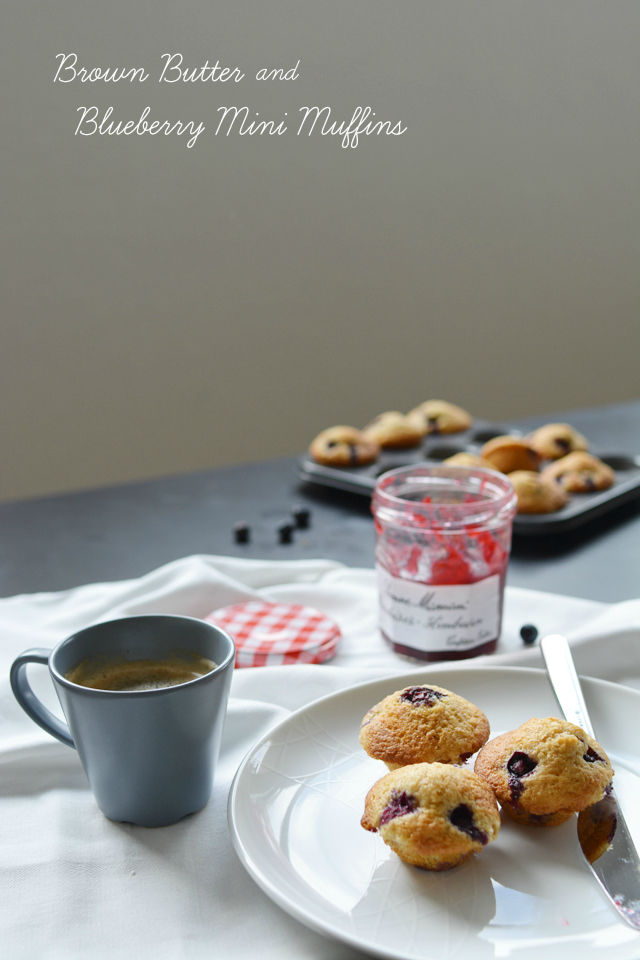 an image of a beautifully arranged table setting with a white plate holding three delectable brown butter and blueberry mini muffins, accompanied by a fork, and a steaming cup of coffee