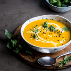 A bowl of turmeric soup with coconut milk drizzled in it. The bowl is on a wooden platter with a smaller bowl of turmeric soup and some herbs.