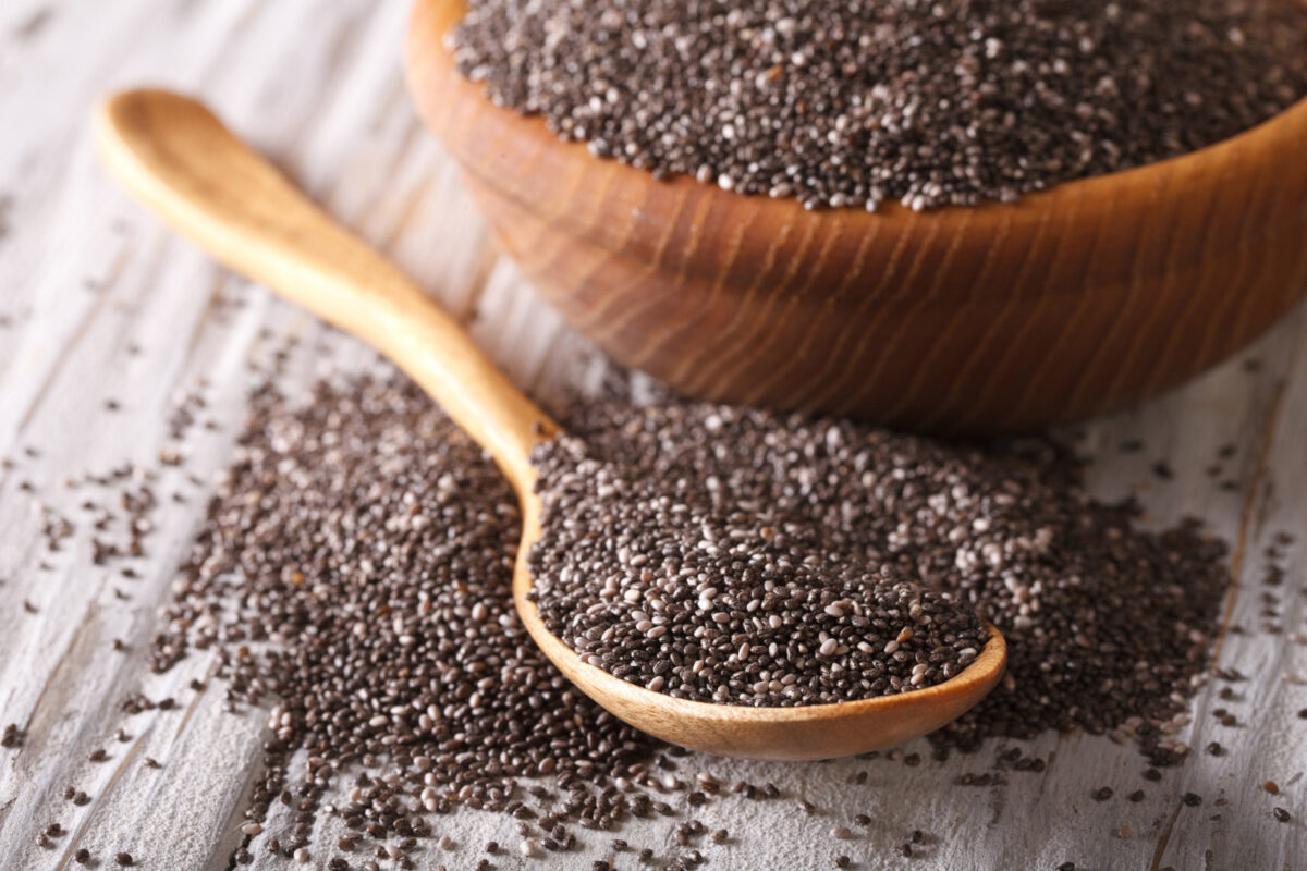 Chia seeds in a wooden spoon on a table with a wooden bowl of chia seeds in the background.