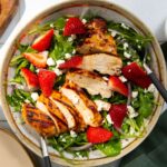 Fresh green salad topped with feta cheese, strawberries, and grilled chicken on a tan and white plate with a white background and a green napkin.