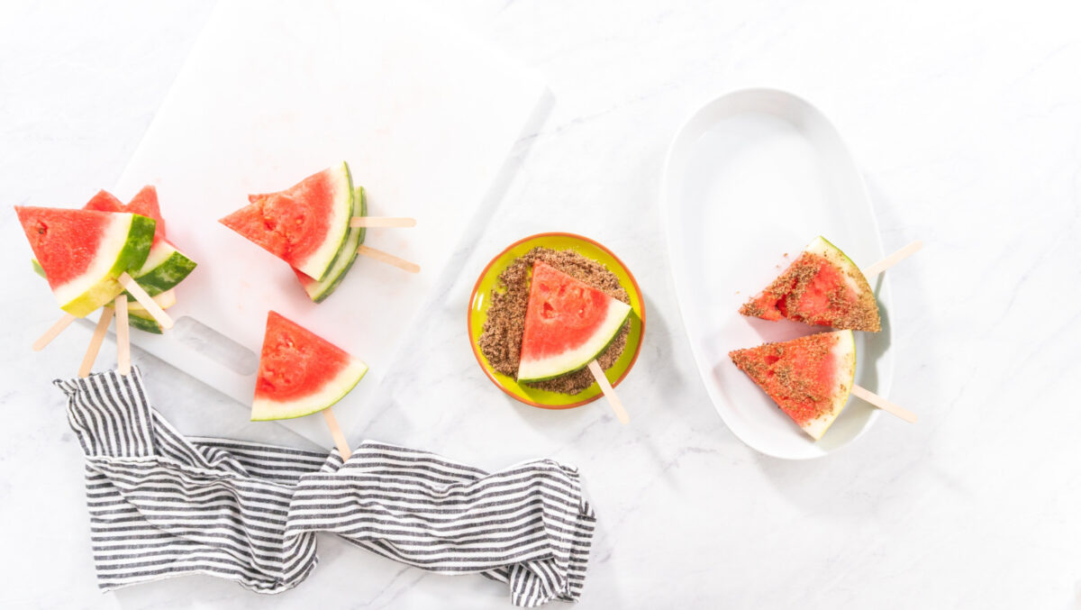 Pieces of watermelon on popsicle sticks being seasoned with Tajin chili lime seasoning in an assembly line fashion.