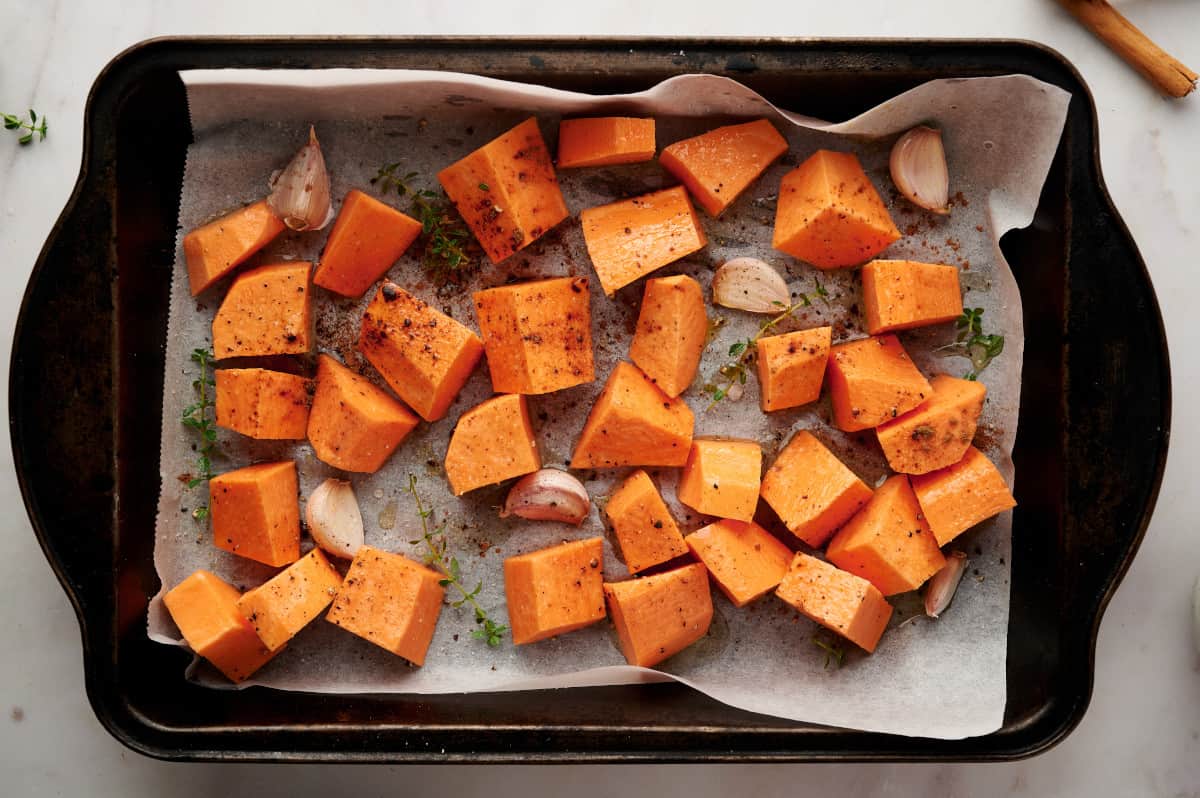 Sweet potatoes and garlic on a parchment lined baking sheet seasoned with thyme and cinnamon.