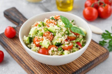 Couscous salad with fresh vegetables and herbs in a bowl on a gray concrete background.