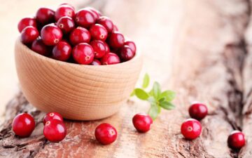 Bright red fresh cranberries in a wooden bowl with some spilled in the foreground on a wood table.