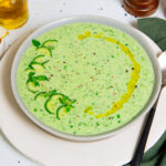 Cold cucumber soup in a white bowl garnished with sliced cucumbers and olive oil.