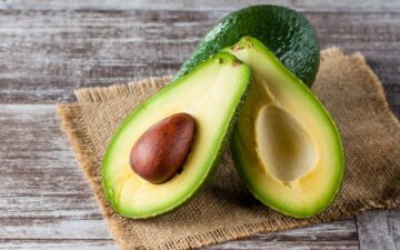 Sliced open avocado with pit propped up on a whole avocado sitting on a piece of burlap on a wooden table.