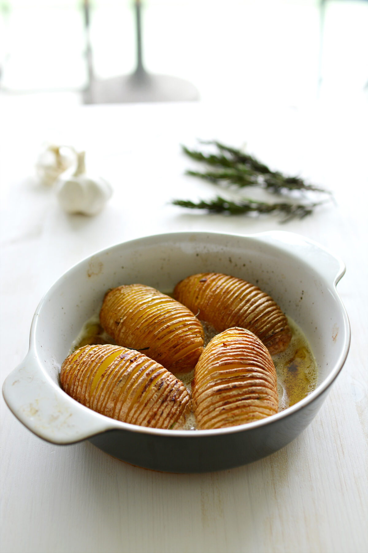 Hasselback Potatoes in a casserole dish on a white table with garlic and rosemary.