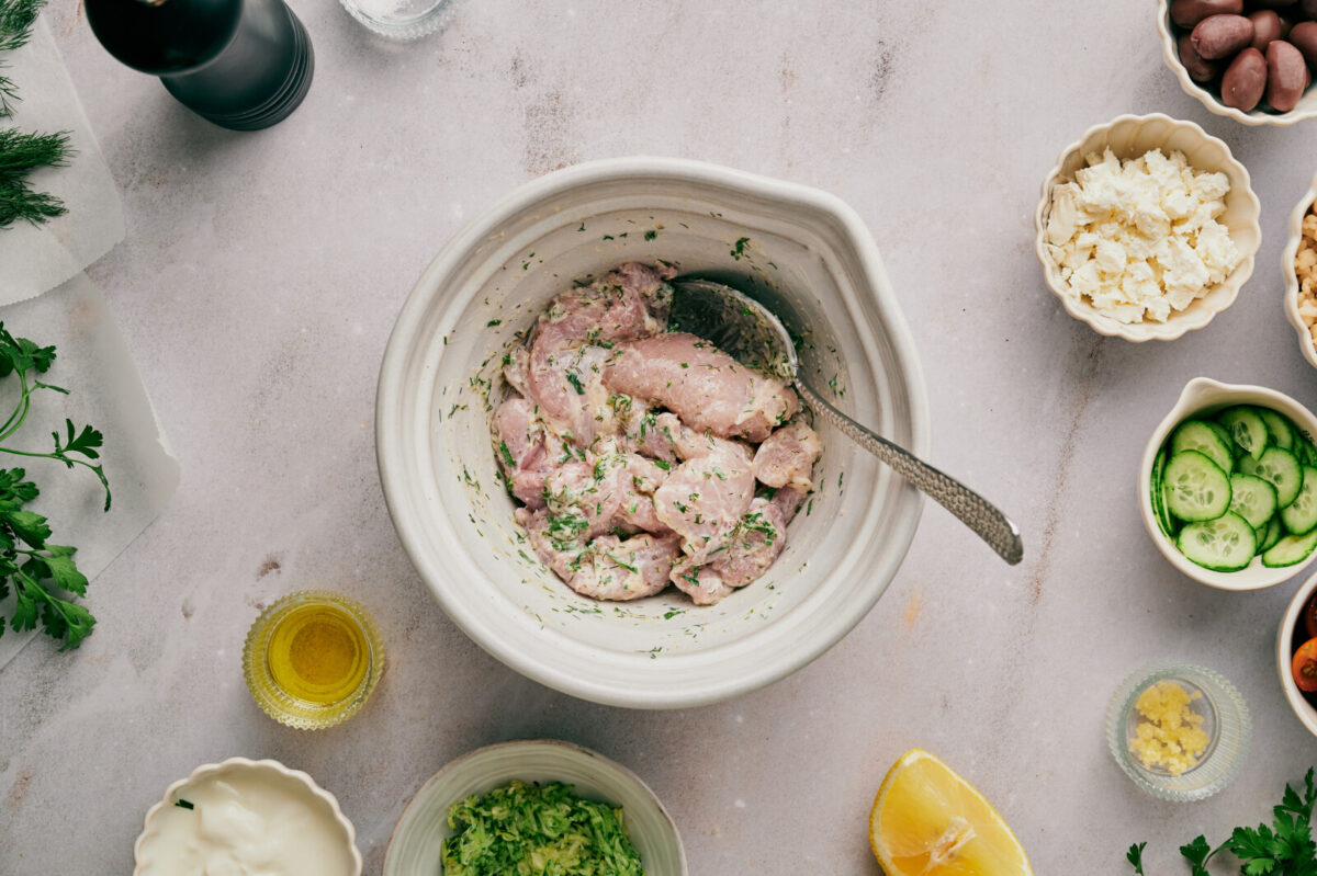 Chicken thighs marinating in a white bowl with smaller bowls of various ingredients.