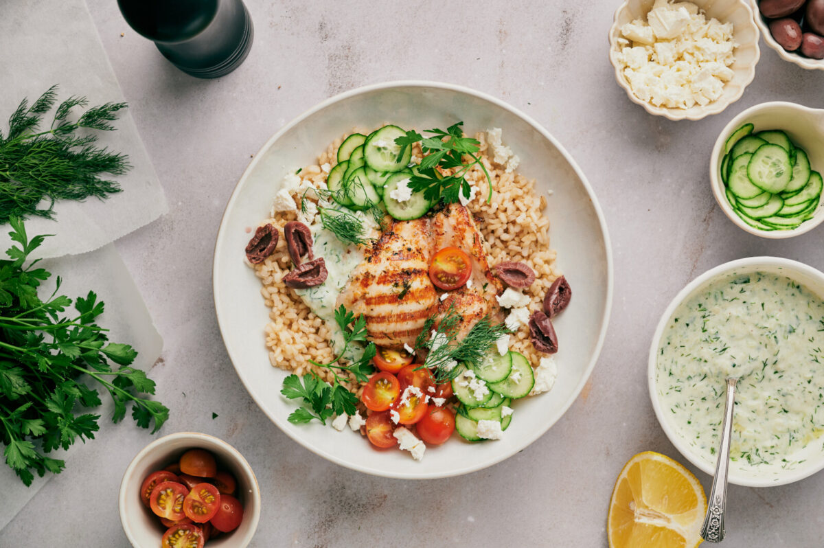 An overhead view of a white bowl filled with rice, Greek chicken, olives, tomatoes, and cucumbers with smaller bowls with various ingredients. 