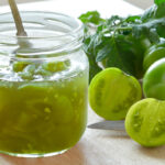 Green tomato chutney in a glass jar with a spoon and green tomatoes in the background.