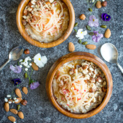 Top-down view of a 2 wooden bowl containing creamy oatmeal adorned with grated apple, toasted hazelnuts, and a sprinkle of sweet brown sugar.