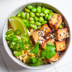 High protein vegetables in a vegan noodle ramen soup with marinated tofu, edamame beans and hot peppers in a gray bowl on a white tile background, top view.