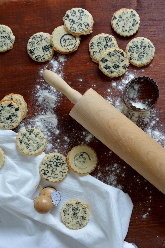 overhead view of stamped marzipan layer cookies scattered on a white cloth dusted with dry flour, accompanied by a rolling pin
