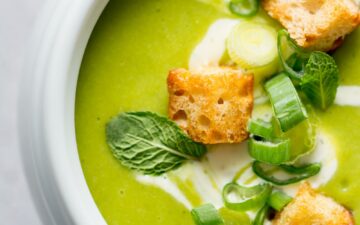 Pea and Mint soup closeup in a white bowl topped with croutons, green onions, and fresh mint leaves.