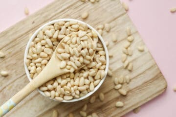 Pine nuts in a white bowl with a wooden spoon on top of a wooden cutting board.