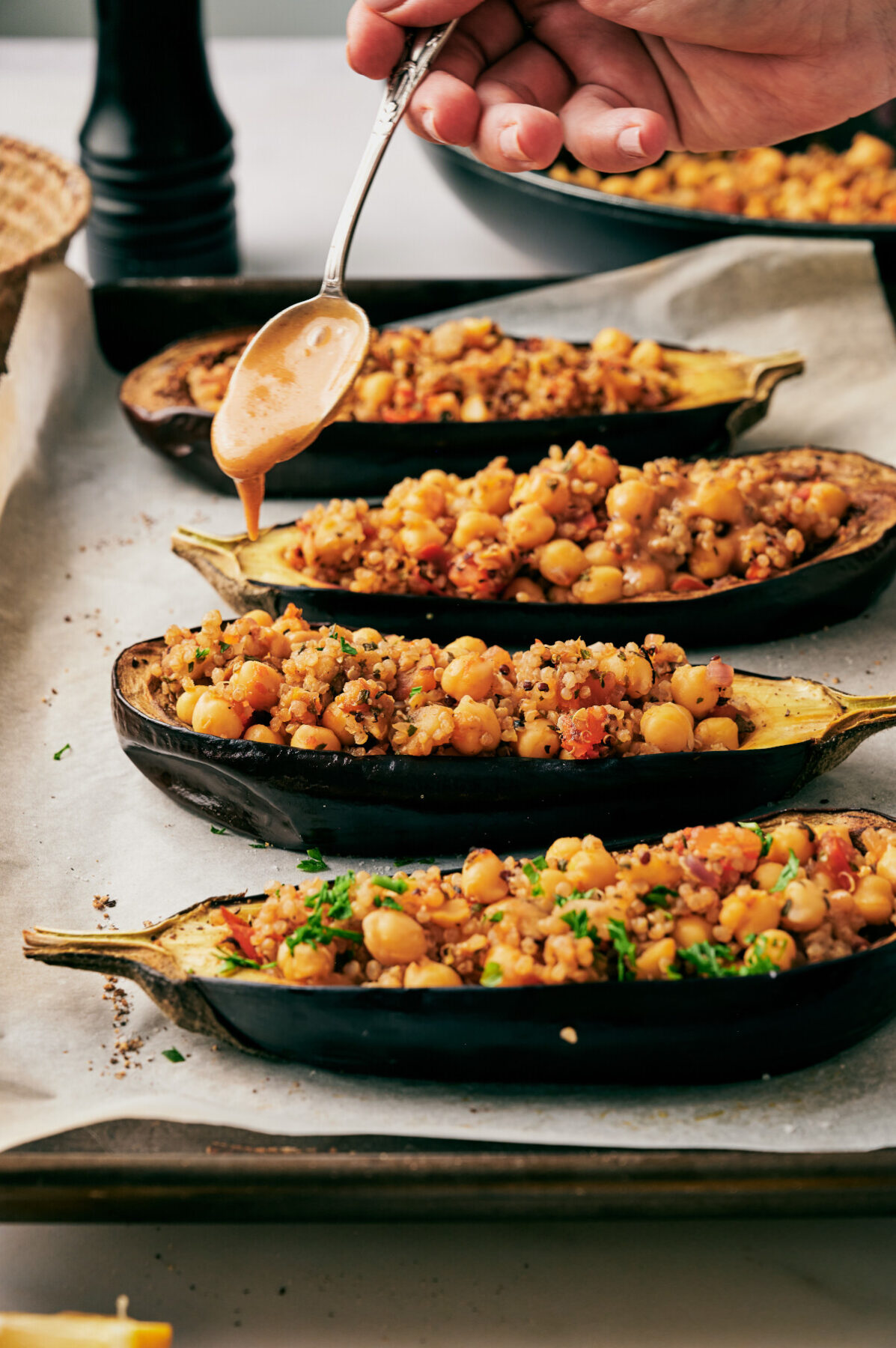 A hand holding a spoon drizzling sauce over quinoa stuffed eggplants on a parchment lined baking sheet.