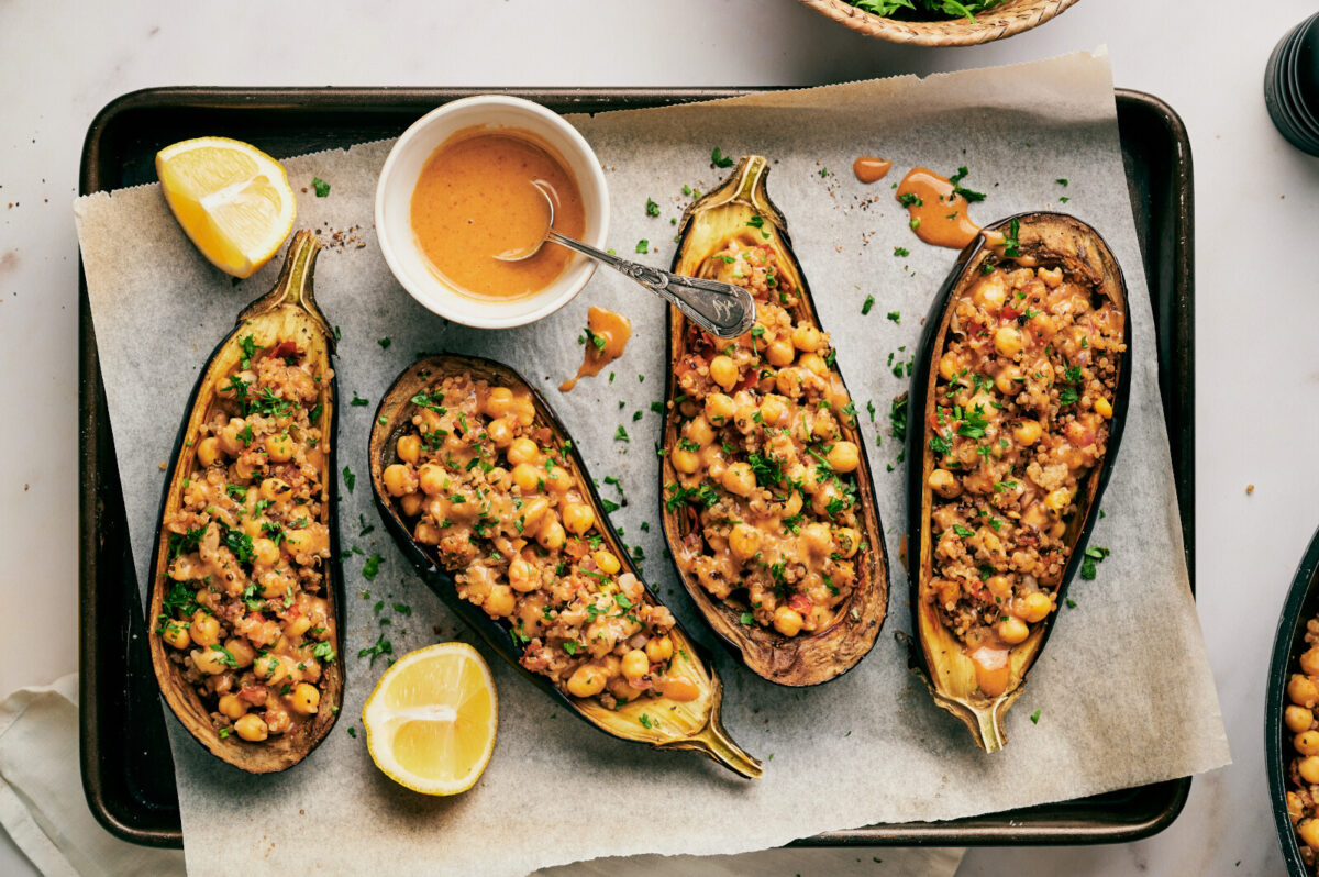 Quinoa Stuffed Eggplants stuffed with chickpea filling on a baking sheet with parchment paper and a ramekin with sauce and a spoon.