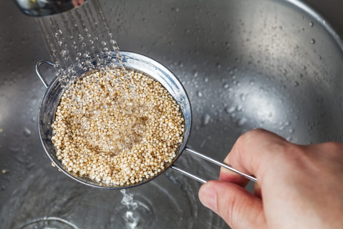 Raw quinoa being rinsed in a metal strainer with a hand holding the strainer.