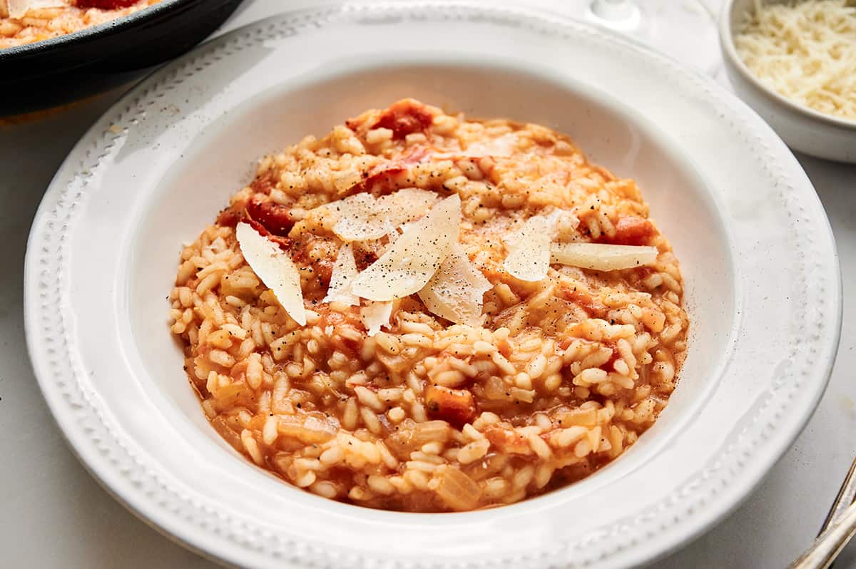 Roasted tomato risotto in a bowl, garnished with fresh Parmesan cheese flakes, salt, and pepper.