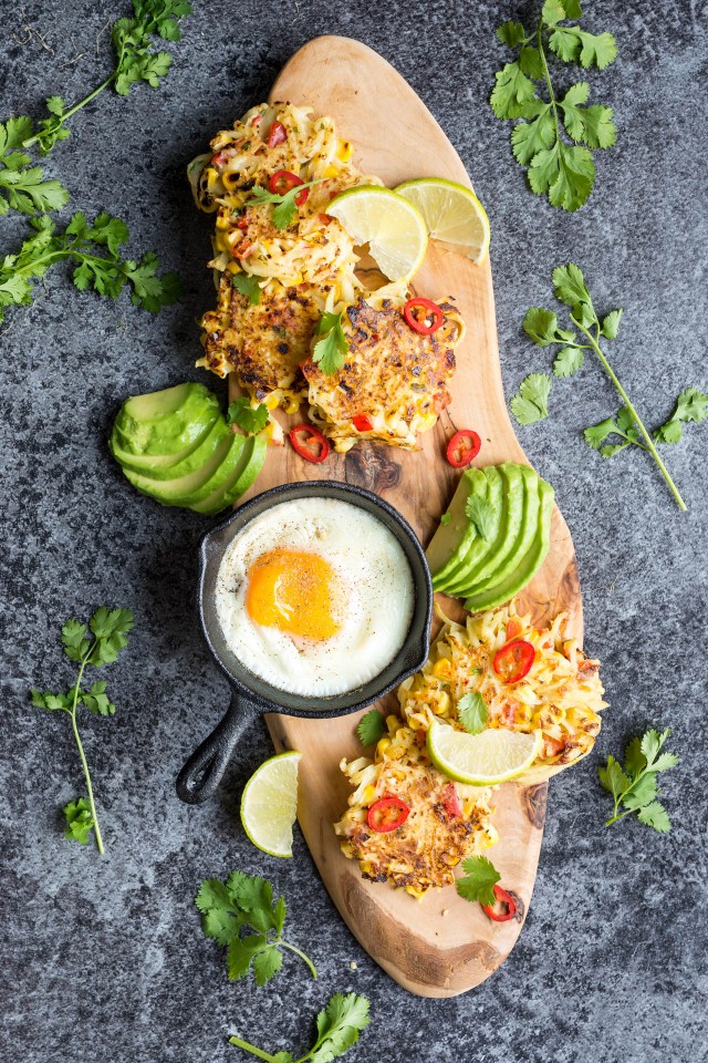 Top-down view of spicy sweetcorn and potato fritters lying flat on a cutting board, accompanied by sliced avocado and a sunny side up egg.