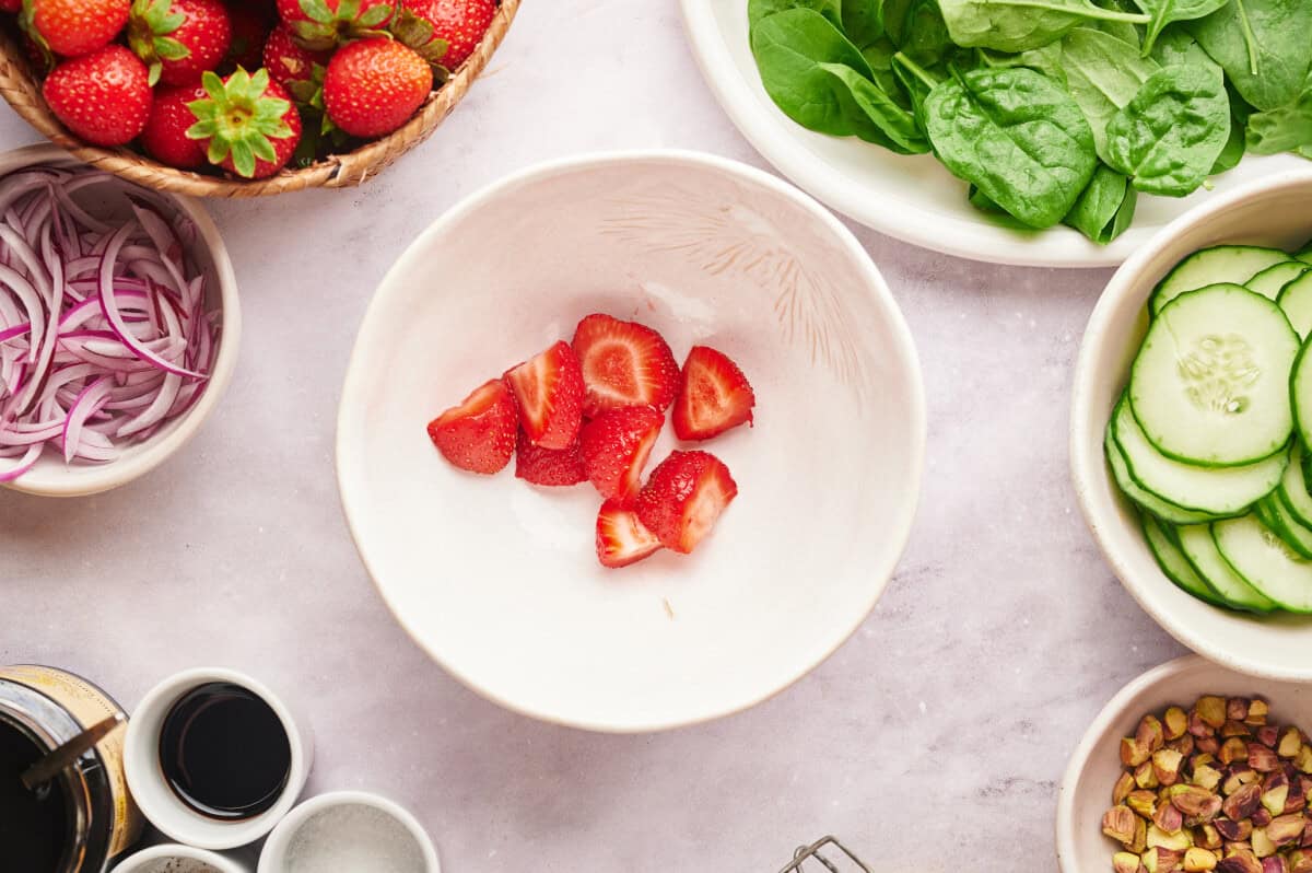 Strawberries cut up in a white bowl, surrounded by various bowls and plates of ingredients to make a strawberry summer salad.