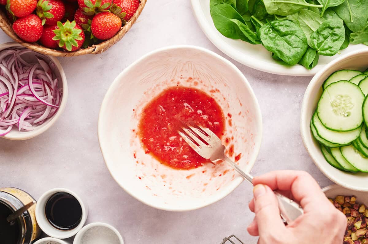 Strawberries being mashed up with a fork in a white bowl, surrounded by various other bowls and plates of ingredients to make a strawberry summer salad.