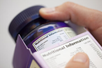 Human hand take out bottle with vitamin pills from the box for reading Nutritional information.