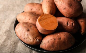 Whole sweet potatoes on a black plate sitting on a burlap tablecloth.