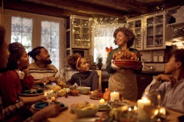 Happy African American mature woman brining stuffed turkey at dining table during family Thanksgiving dinner.