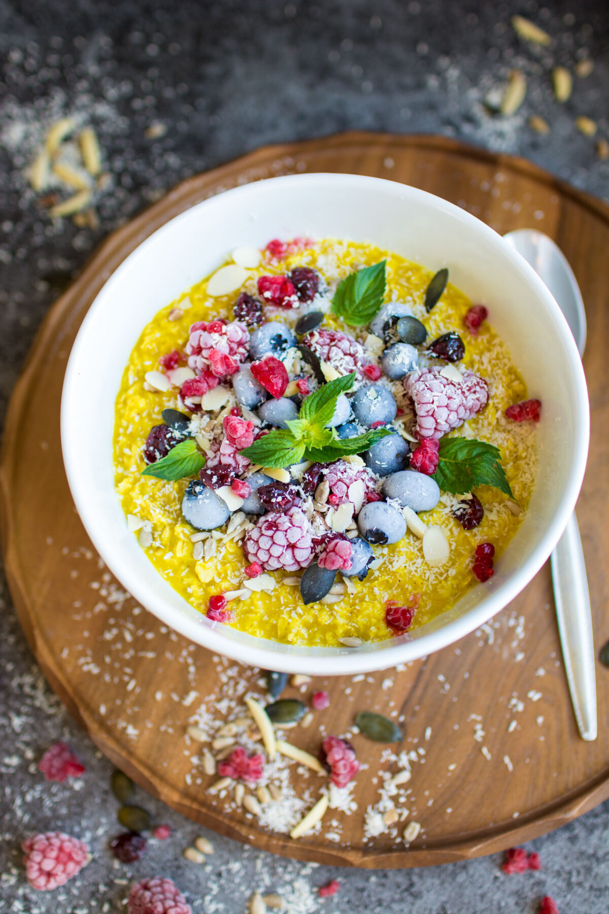 A bowl of golden turmeric oatmeal topped with raspberries, blueberries and mint on a wooden cutting board. 