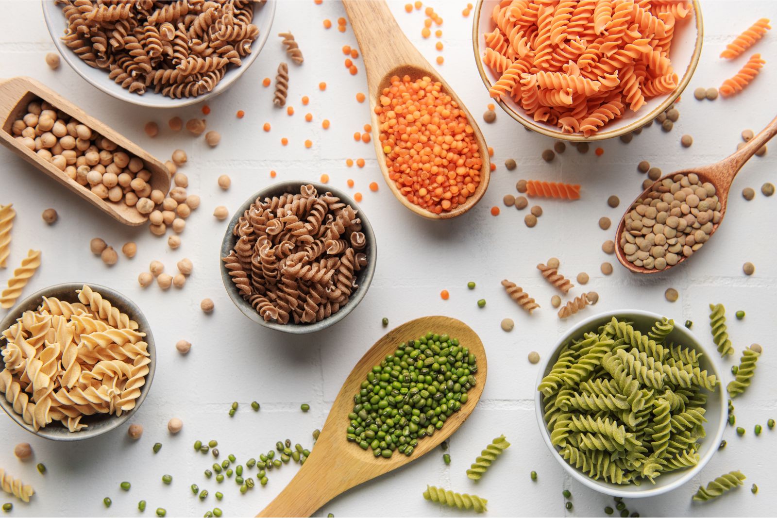 Various flavors of gluten free pasta in small bowls pictured next to large spoons of various legumes on a white background.