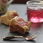 Violet jelly spread on a biscuit with a spoon in front and a jar of violet jelly in the background.